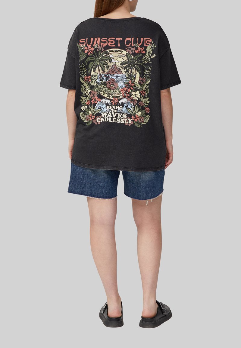 Zwarte oversized t-shirt met een kleurrijk graphic ontwerp van tropische elementen, tekst en bloemen op de achterkant, gecombineerd met spijkerbroeken.