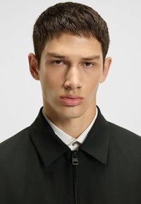 Black jacket with a smooth texture, front zipper, and a collared design, paired with a white shirt visible at the collar.