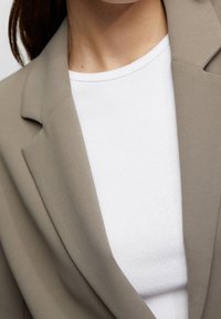 Blazer brun clair à texture lisse, avec des revers structurés. En dessous, un haut blanc côtelé mettant en valeur les couleurs contrastées.