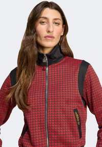 Röd och svart zip-up jacka med ett hundtandsmönster, hög krage och svarta axelaccenter. Inkluderar en silverfärgad adidas-logotag.