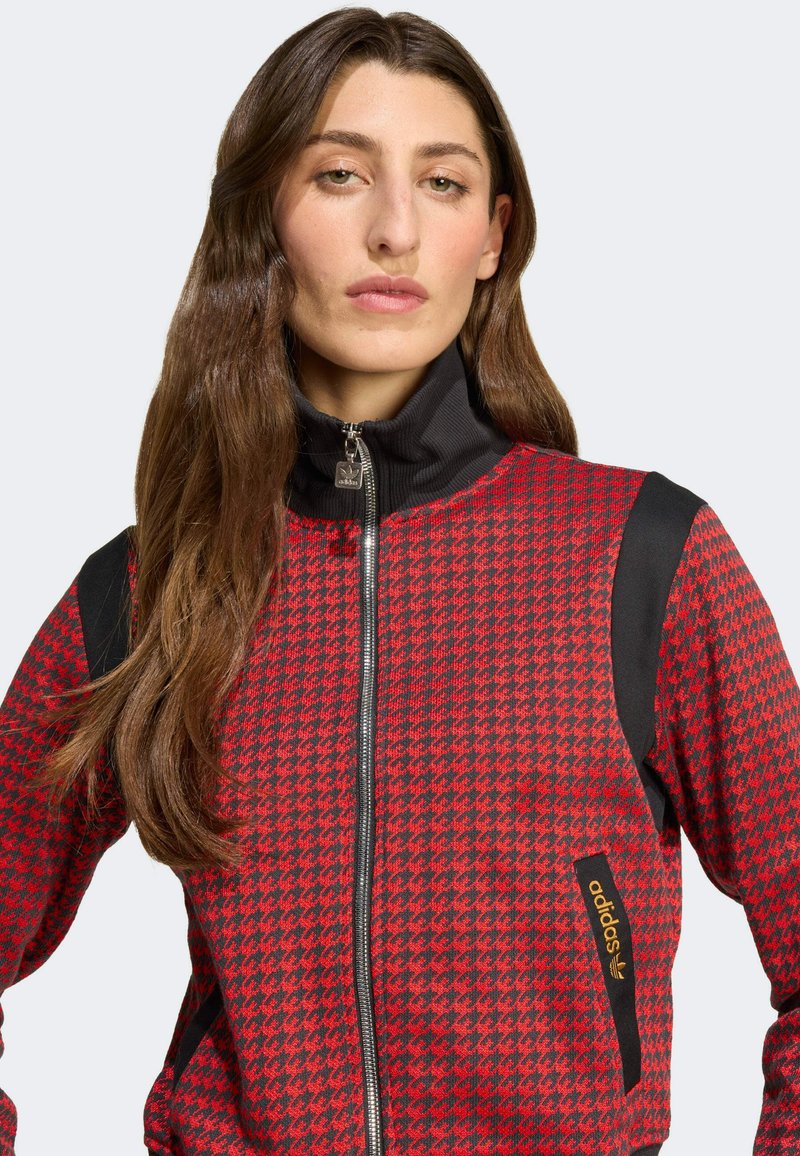 Giacca zip-up rossa e nera con motivo a zig zag, colletto alto e dettagli neri sulle spalle. Include un'etichetta del logo adidas argentata.
