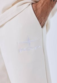 Vêtement couleur crème avec une texture douce, arborant un logo brodé blanc "PXP" et un symbole d'étoile décoratif sur le côté.
