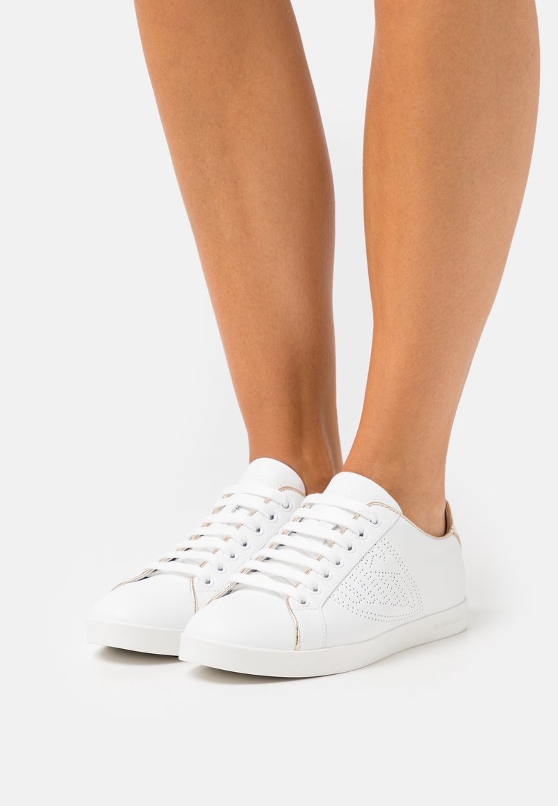 Sneakers bianche a taglio basso con una superficie in pelle liscia, design perforato sul lato, accenti beige e lacci bianchi. Stile minimalista.