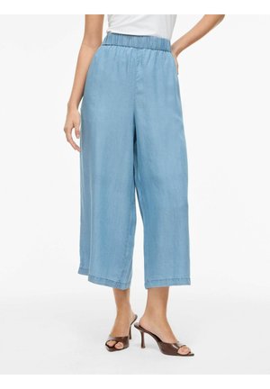 Kvinde iført lyseblå viddebukser med elastik i taljen og brune hælede sandaler med åben tå, stående på hvid baggrund.