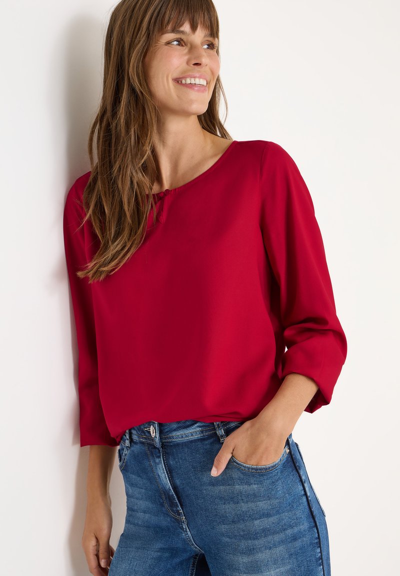 Rød bluse med trekvartærmer, rund hals og knapdetalje. Kombineret med lyseblå denimjeans med sidelommer.