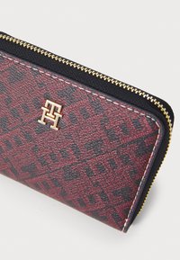 Red and black patterned wallet with a textured surface, gold hardware, and a zip closure. Features a prominent logo in gold.