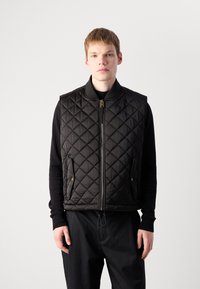 Black quilted vest featuring a zip closure, side pockets with gold hardware, and a ribbed collar. Worn over a black long-sleeve shirt.