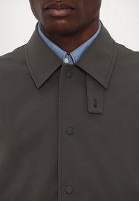 Homme portant un manteau gris foncé boutonné par-dessus une chemise bleu clair à col, visible du cou jusqu'en haut de la poitrine.
