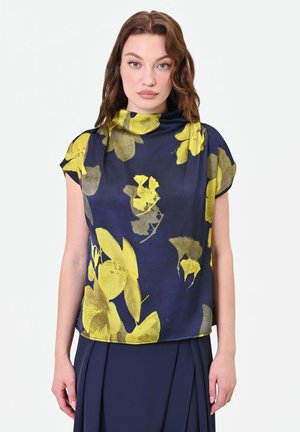 Donna che indossa un top blu navy con grandi motivi floreali gialli e collo alto, abbinato a una gonna blu navy scuro.
