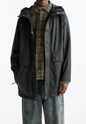 Osoba ubrana w rozpiętą czarną kurtkę przeciwdeszczową z kapturem, na zielonej, w kratę koszuli na guziki i luźnych niebieskich dżinsach, z pierścieniami na prawej dłoni.