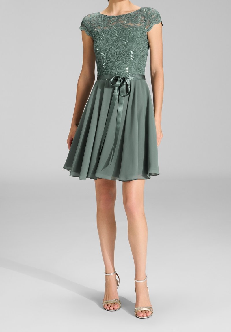 Femme portant une robe courte en dentelle verte avec un ruban de satin à la taille et une jupe plissée, associée à des sandales à talons hauts argentées.