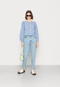 Blusa de mangas longas azul com padrão floral, calças de ganga de cintura alta xadrez azul claro, sandálias brancas com tiras e uma pequena mala verde.