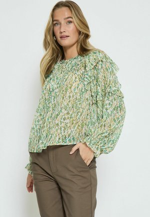Blumenbluse in Creme mit grünen und blauen Mustern, ausgestattet mit Rüschensäumen und einer lockeren Passform, kombiniert mit braunen Hosen.