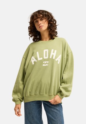 Frau mit lockigem Haar, die einen lockeren hellgrünen Sweatshirt mit dem Text "ALOHA" und blaue Jeans trägt, vor einem schlichten Hintergrund.