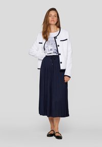 Weißer strukturierter Blazer mit marineblauen Akzenten und Knöpfen, kombiniert mit einem grauen T-Shirt mit Grafik und einem marineblauen Faltenrock in Midi-Länge. Schwarze flache Schuhe.