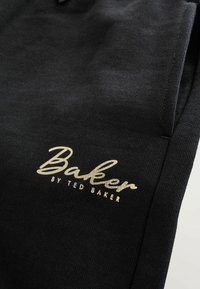 Joggers de tela negra con textura sutil. Presentan el logo dorado "Baker by Ted Baker". Incluyen bolsillos laterales y un diseño de corte relajado.