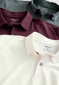 Három pólóing, sötétzöld, bíborvörös és fehér színben, klasszikus gallérral és gombos felsőrész kialakítással. A címkék "NEXT" és "REGULAR FIT" feliratot viselnek.