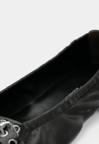 Ballerine en cuir noir avec une texture lisse, un bout arrondi et un détail de perforation décoratif sur le dessus. L'intérieur présente un revêtement sombre.