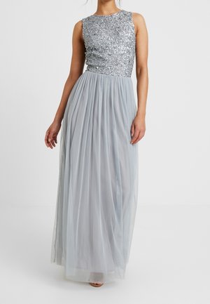 Vrouw draagt een mouwloos zilveren top met pailletten en een vloeiende, lichtgrijze, geplooide rok tot op de vloer, staand tegen een effen achtergrond.