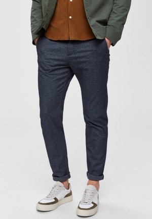 Man wearing navy blue slim pants, rolled cuffs, white and olive green sneakers, brown shirt, and green jacket with hands in pockets.