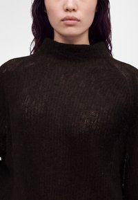 Gros plan d'une personne portant un pull en maille noire duveteuse avec un col rond haut sur un fond uni.