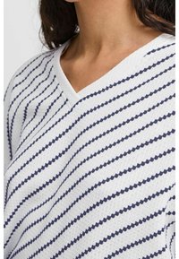 Primo piano di una persona che indossa un maglione bianco a V con strisce diagonali a pois blu navy.