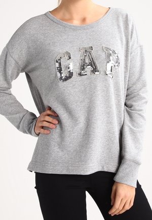 Vrouw draagt een lichtgrijs sweatshirt met lange mouwen en grote zilveren pailletten met "GAP" op de borst, gecombineerd met een zwarte broek.