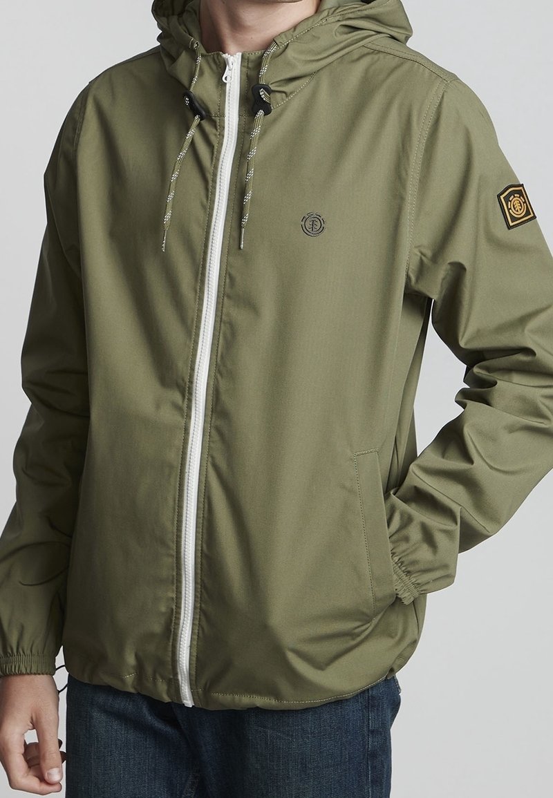 Veste vert olive avec une fermeture éclair blanche à l'avant, capuche ajustable avec cordon, logo sur la poitrine et un patch sur la manche. Comprend des poches latérales.