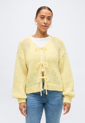 Femme portant un cardigan tricoté jaune pâle noué avec trois nœuds sur un haut blanc et un jean bleu, debout devant un fond uni.