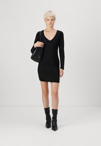 Vestido preto de mangas longas com decote em V, feito de tecido texturizado. Combinado com uma bolsa preta de ombro e botas pretas até ao joelho.