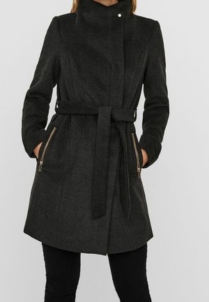 Woman wearing a dark gray belted coat with a high collar and zippered pockets, paired with black pants, standing with hands in pockets.
