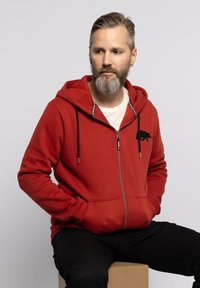 Felpa rossa con zip, realizzata in tessuto morbido, presenta una silhouette di orso nero sul petto, cappuccio con cordino regolabile e tasche frontali.