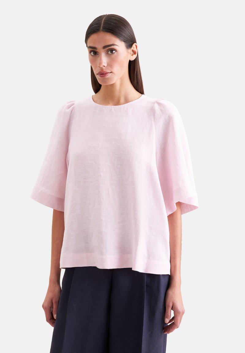 Femme aux cheveux lisses et foncés porte un chemisier ample rose clair avec des manches bouffantes mi-longues et un pantalon large foncé sur un fond uni.