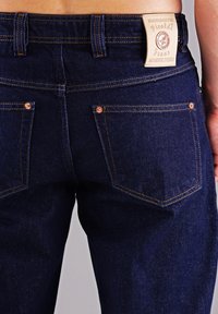 Dunkelblaue Jeans aus Denim mit glatter Textur. Verfügt über ein Etikett am hinteren Bund und zwei aufgesetzte Gesäßtaschen mit kupferfarbenen Nieten.