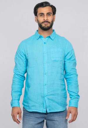 Homme barbu portant une chemise bleu vif à manches longues boutonnée et un jean bleu, debout devant un fond gris uni.