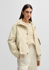 Jachetă bej scurtă cu închidere prin capse, guler mare și buzunare laterale, purtată peste un pulover texturat crem și pantaloni bej deschis.