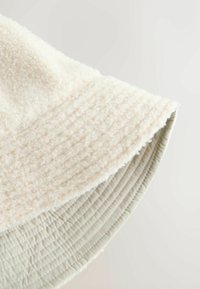 Soft cream bucket hat with textured fleece fabric and smooth, quilted brim. Rounded shape with subtle stitching detail along the edge.