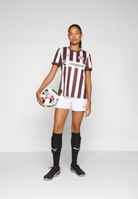 Brown and white striped soccer jersey with logo, white shorts, black socks, and black cleats. Player holds a black and white soccer ball.