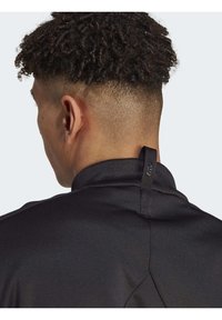 Chaqueta deportiva negra con un cuello alto, tejido texturizado y una pequeña etiqueta en forma de lazo en la parte posterior. Piel visible en el cuello y la oreja.