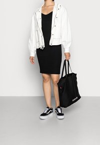 Giacca bianca con zip, vestito nero a coste, sneakers nere con suola bianca, che porta una borsa a tracolla nera Levi's. Texture lisce e colori neutri.
