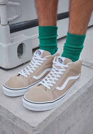 Piernas con calcetines verdes y zapatillas Vans altas beige con cordones blancos y franjas laterales, de pie sobre una superficie de hormigón.