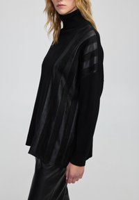 Kobieta w czarnej bluzie z golfem z skórzanymi wstawkami z przodu i na rękawach, w połączeniu z czarnymi skórzanymi spodniami.