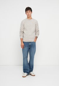 Homme portant un pull en tricot gris clair, un jean bleu et des baskets blanches, se tenant avec une main dans la poche contre un fond blanc uni.