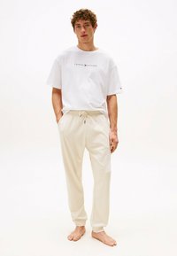 Camiseta blanca de manga corta con el logo "TOMMY HILFIGER" y pantalones de chándal beige con cordones y puños ajustados. El modelo está descalzo.