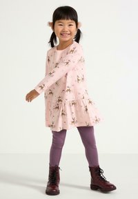Rosa langærmet kjole med hjorte- og blomsterdesign, klokket nederst, parret med lilla leggings og sorte snørebåndsstøvler.