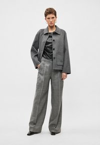 Γκρι μάλλινο σακάκι με γιακά και μπροστινή τσέπη, συνδυασμένο με γκρι καρό παντελόνι φαρδιάς γραμμής και μαύρο γυαλιστερό τοπ. Μαύρα παπούτσια.