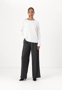 Blusa bianca a maniche lunghe con dettagli arricciti ai polsi, abbinata a pantaloni larghi grigi scuri a vita alta e scarpe nere slip-on.