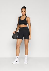 Czarny sportowy biustonosz i zestaw szortów o dopasowanym kroju, gładkiej fakturze i z logo. Modelka trzyma czarną torbę, ma na sobie białe sneakersy.