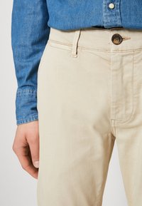 Light beige trousers with a subtle texture, featuring a button closure and side pockets. Worn with a blue denim shirt.