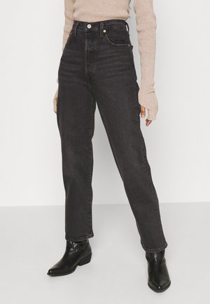 Person wearing high-waisted black straight-leg jeans, beige ribbed sweater with thumb holes, and black ankle boots, standing against a gray background.